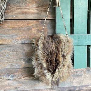 May include: A small, rectangular bag made of brown and tan fur, suspended by a braided cord. The bag hangs against a weathered wooden wall and a green painted wooden structure.