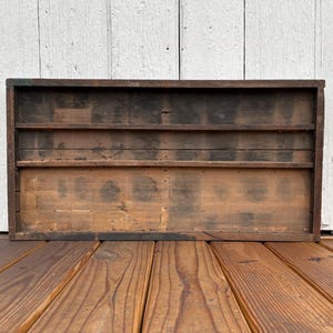 May include: A rectangular, wooden display shelf with two interior shelves. The wood has a distressed, aged appearance with dark and light brown tones. The shelf is set against a white wooden background.