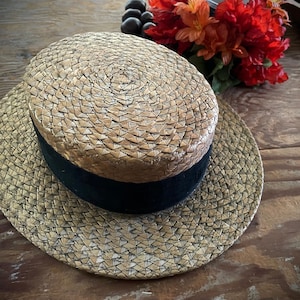 May include: A straw boater hat with a black band. The hat has a wide brim and a flat top. The hat is made of woven straw and is a light tan color. The hat is sitting on a wooden surface.
