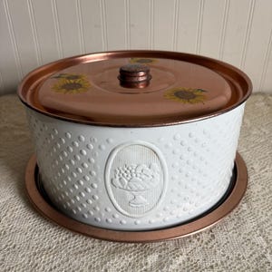May include: White metal bread box with a copper-colored lid and base. The lid features yellow sunflower designs. The box has a textured pattern and an oval emblem with a fruit bowl design. A vintage kitchen storage item.