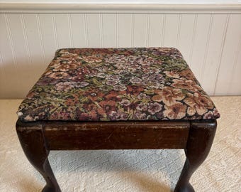May include: A square footstool featuring a dark brown wooden frame with four curved legs and a padded seat upholstered in a multi-colored floral tapestry fabric with shades of red, pink, purple, and green flowers on a dark background.