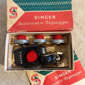 May include: Vintage Singer Automatic Zigzagger sewing machine attachment in its original box. The box is red, white, and teal with the Singer logo. The attachment is black with red accents and silver knobs. The box also contains a manual.