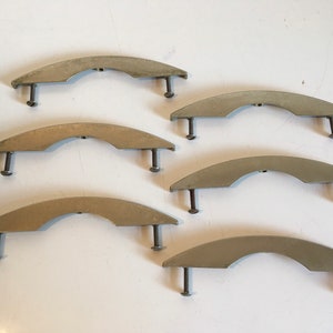 May include: Six silver-toned, curved cabinet handles with attached screws. The handles have a semi-circular design and are arranged on a white surface. The metal has a slightly aged appearance.
