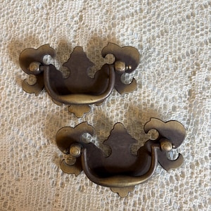 May include: Two antique brass drawer pulls. Each pull features a curved handle and decorative floral-like accents. The pulls have a dark brown finish with gold highlights, and are set against a cream-colored, textured background.