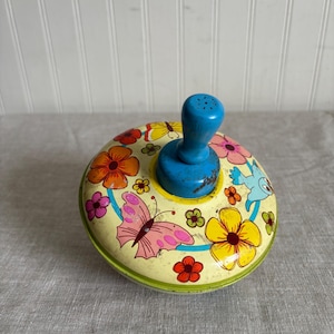 May include: Vintage metal spinning top with a blue handle. The top is decorated with colorful flowers, butterflies, and a blue bird on a pale yellow background. The top has a green rim.