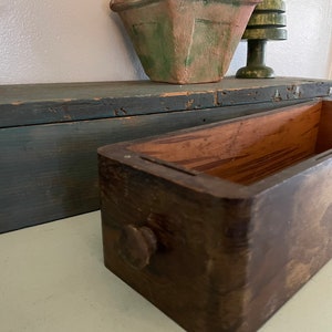 May include: A weathered wooden drawer with a round knob, set against a backdrop of a teal-painted wooden box and a terracotta-colored planter with greenery. A green decorative tree is also visible.