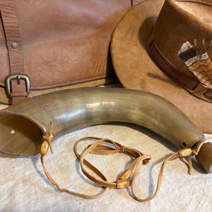 May include: A curved, horn-shaped container with a leather strap, likely a powder horn. The horn is a light brown color with a polished finish. A brown leather satchel and a brown hat with a feather are in the background.