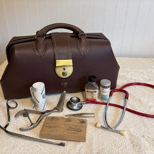 May include: A dark brown leather medical bag with a brass latch and the initials J.H. embossed on the front. Included are a red stethoscope, metal surgical instruments, a roll of white medical tape, two small glass medicine bottles, and a box.