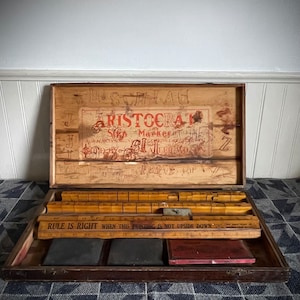 May include: Vintage wooden printing set in an open box. The box contains wooden letter blocks, rulers, ink pads, and the text "ARISTOCRAT Marker". The box is on a patterned fabric.