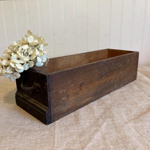 May include: A rectangular, dark brown wooden box with a handle on the front. The box is empty and has a distressed finish. A bundle of dried, cream-colored flowers rests on the edge of the box. The box is likely used for storage or display.