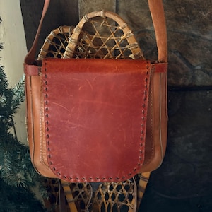 May include: A handcrafted leather bag with a red flap and a long strap. The bag is filled with a pair of vintage snowshoes. The bag is brown with visible stitching and a rustic aesthetic.