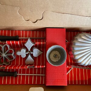 May include: A vintage rosette iron set in a cardboard box. The set includes various shaped irons, such as a flower, diamond, and heart, with black handles. The box has a red and white striped interior.