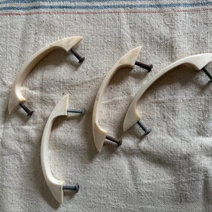 May include: Four off-white, curved cabinet handles with attached screws. The handles have a smooth, slightly glossy finish and are arranged on a textured, light-colored fabric. The screws are a dark, metallic color.