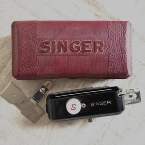 May include: A vintage Singer sewing machine accessory kit. The kit includes a burgundy case with "SINGER" embossed on the lid, a black sewing machine attachment, and several metal pieces. The background is a light, textured surface.