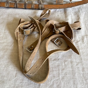 May include: Close-up of vintage snowshoe bindings made of light brown leather. The straps have metal buckles and are arranged in a tangled configuration. The background shows a portion of a wooden snowshoe with a woven design.