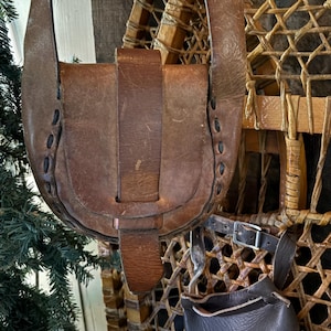 May include: A brown leather satchel with a flap closure and a long strap hangs in front of a pair of vintage snowshoes. The satchel has decorative stitching and a buckle closure. The snowshoes are made of wood and woven material.