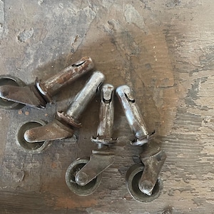 May include: A collection of five antique metal casters, showing signs of age and wear. Each caster has a wheel and a stem, with a rusty, aged appearance. The casters are arranged on a weathered wooden surface.