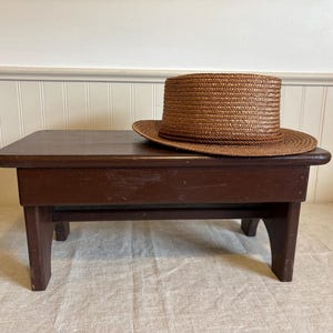 May include: A small, dark brown wooden step stool with four legs. A brown straw hat rests on the stool's surface. The hat has a wide brim and a cylindrical crown. The stool is placed on a light-colored fabric.