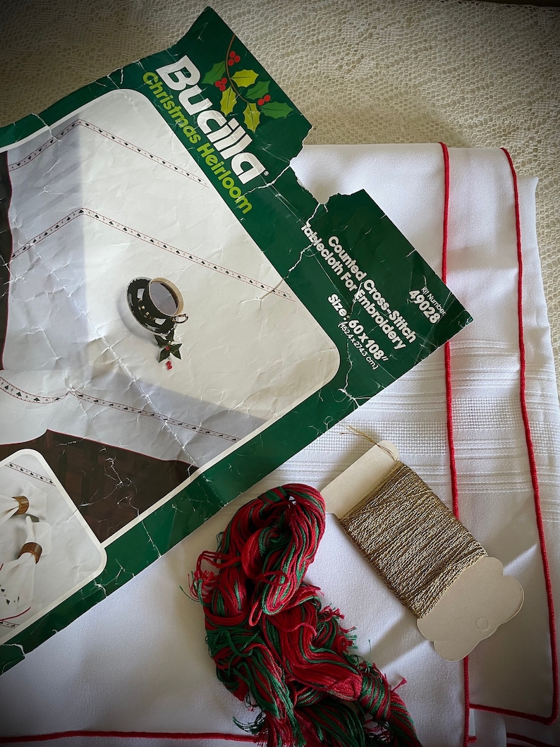 K&ouml;nnte beinhalten: Ein Bucilla Christmas Heirloom Kreuzstich-Tischdecken-Set. Das Set enth&auml;lt eine wei&szlig;e Tischdecke mit roter Bord&uuml;re, Stickgarn in Rot und Gr&uuml;n sowie eine Spule Goldfaden. Die Tischdecke misst 152,4 x 274,3 cm.
