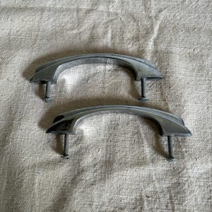 May include: Two vintage, curved metal cabinet handles with attached screws. The handles are silver-toned and have a slightly arched design. They are likely for a drawer or cabinet door.