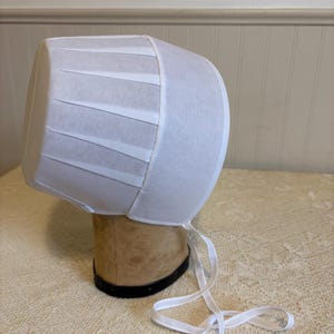 May include: A white bonnet with a pleated design and a ribbon tie. The bonnet is made of a lightweight fabric and is displayed on a mannequin head. The bonnet is a vintage style.