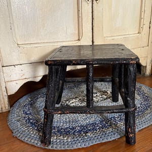 May include: A small, rustic, black wooden stool with four legs and a rectangular seat. The stool is placed on a blue and white braided rug. The background features a white cabinet with blue knobs.