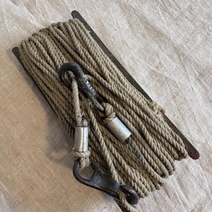 May include: A coiled rope with metal hardware, including a hook and cylindrical connectors. The rope is a neutral beige color, and the metal components have a weathered, antique appearance. The rope is wound around a dark metal spool.