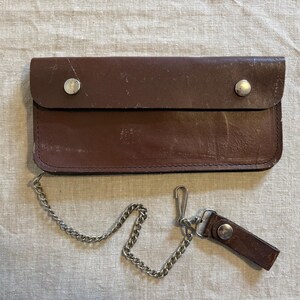 May include: A rectangular brown leather wallet featuring a fold-over flap secured by two silver-toned metal snaps. A silver-toned metal chain is attached to the side, ending in a small leather tab with a matching snap closure.
