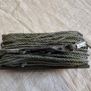 May include: A coiled, olive-green rope with a black metal clasp and silver metal ends. The rope is tightly wound and rests on a dark, rectangular base. The clasp has text on it.