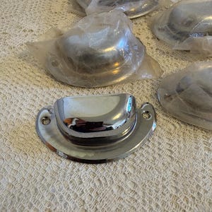 May include: A collection of polished chrome cup pulls, some still in their original plastic packaging. The pulls have a semi-circular design with a rounded cup and a flat base with two screw holes. The background is a textured, off-white surface.