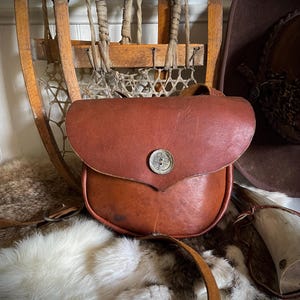 May include: A brown leather pouch with a rounded flap and a silver button closure. The pouch is set against a backdrop of vintage snowshoes, a cowboy hat, and a fur rug, evoking a rustic, outdoorsy aesthetic.