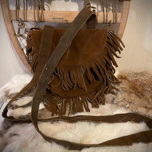 May include: A brown suede shoulder bag with a flap and fringe detail. The bag has a long strap and is set against a backdrop of a fur rug and a wooden chair.