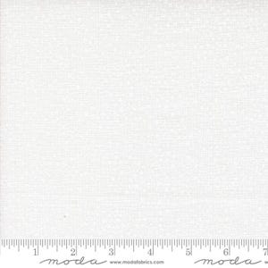 May include: A solid white fabric with a subtle, fine-textured weave pattern. A ruler is visible along the bottom edge, showing measurements in inches, with the text "moda" and "www.modafabrics.com" printed repeatedly along the selvage edge.