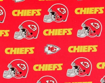 May include: A red fabric featuring a repeating pattern of Kansas City Chiefs football helmets in red and white, the KC arrowhead logo, and the word CHIEFS in yellow text. A white ruler with black markings is visible along the bottom edge.