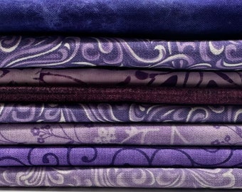 May include: A stack of various purple fabric swatches. The fabrics include solid colors, swirls, and floral patterns. The shades of purple range from deep indigo to light lavender. The fabrics are neatly stacked, showcasing the different textures and designs.