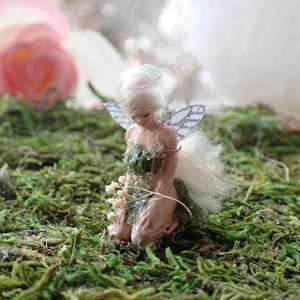 May include: A miniature fairy figurine with white hair and delicate, patterned wings. The fairy is kneeling on a bed of green moss, holding a small bouquet of white flowers. The figurine is painted in soft, natural tones.