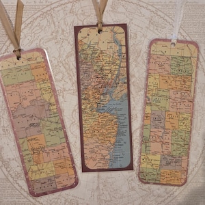 May include: Three rectangular bookmarks featuring vintage map prints in muted tones of yellow, green, and pink. Each bookmark has a hole at the top with a ribbon attached. The center bookmark displays a map of New Jersey, while the others show county maps.