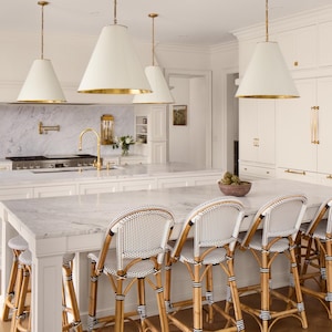 May include: A white kitchen with a large island and white cabinets. The island has a white marble countertop and four white and black wicker bar stools. There are two gold pendant lights hanging over the island.