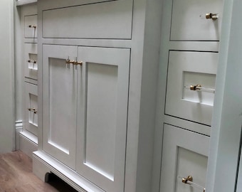 May include: White bathroom vanity with two cabinet doors and three drawers. The cabinet doors have gold-toned hardware. The drawers have clear acrylic handles with gold-toned accents.