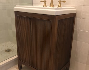 May include: A brown wooden bathroom vanity with a white porcelain sink and gold faucets. The vanity has a fluted design and sits on four legs.