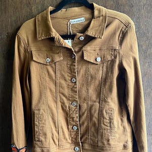 May include: A brown denim jacket featuring a classic collar, a button-up front with silver-toned metal buttons, two chest pockets with flaps, and two side pockets. A colourful butterfly patch is visible on the lower right sleeve.