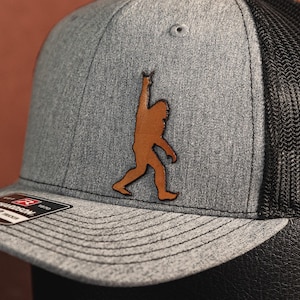May include: A gray and black trucker hat with a brown leather patch of a Bigfoot silhouette. The patch shows Bigfoot with one arm raised in the air.