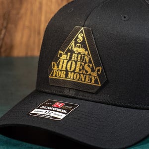 以下が含まれることがあります： 三角形の金色のパッチが付いた黒いトラッカーハット。パッチには「I run hoes for money」と書いてあります。