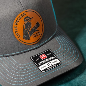 May include: A gray and blue baseball cap with a brown leather patch featuring a woodpecker and the text "Little Pecker Club". The cap also has a black and white label with the text "Est. 1970 Richardson Original 112 Trucker Adjustable".