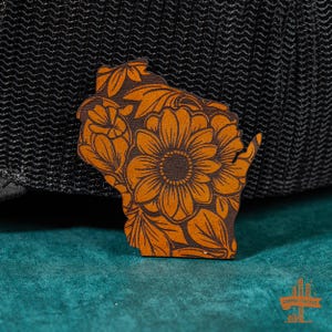 May include: A brown leather cutout of the state of Wisconsin with a floral design. The design features a large sunflower in the center surrounded by smaller flowers and leaves.