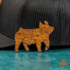 May include: A brown leather pig patch with a floral design. The patch is attached to a black mesh trucker hat.