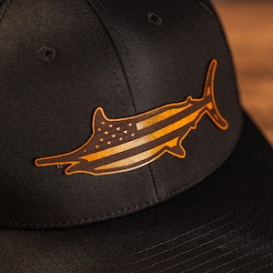 May include: A black baseball cap with a gold marlin silhouette featuring an American flag design.