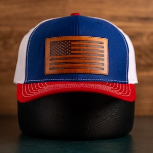 Op de afbeelding: Een blauwe en witte truckercap met een rode rand. De pet heeft een bruin leren Amerikaans vlagpatch.