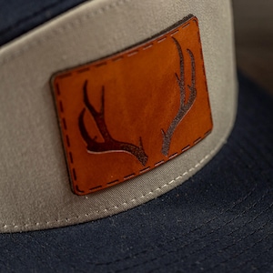 May include: A beige and navy blue baseball cap with a brown leather patch featuring a stylized antler design.