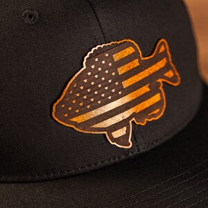 May include: A black baseball cap with a brown leather patch shaped like a fish with an American flag design.